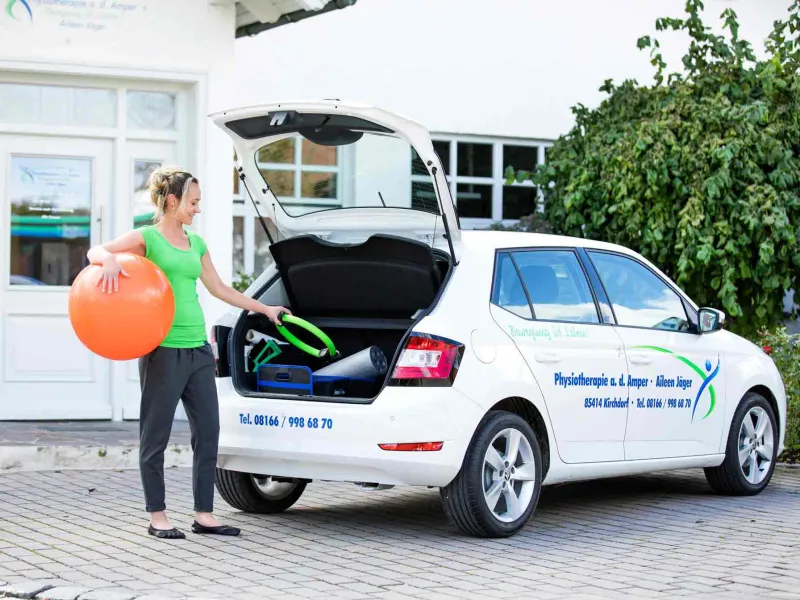 Physiotherapeutin lädt einen Gymnastikball in das beschriftete Praxisauto